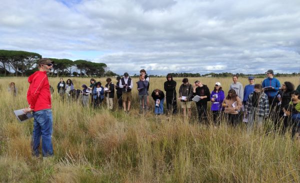 One person addresses a line of students standing in a grassy paddock.