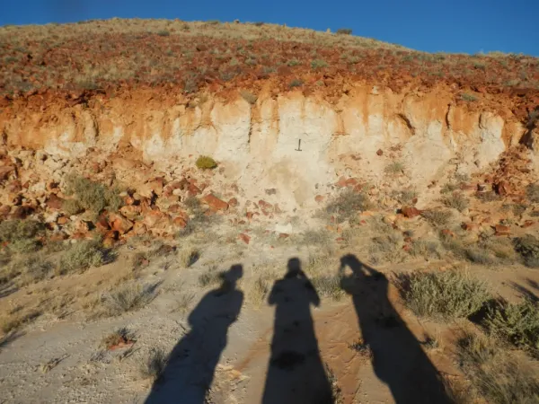 Three shadows of people falling against the side of a hill, with sediment layers visible.
