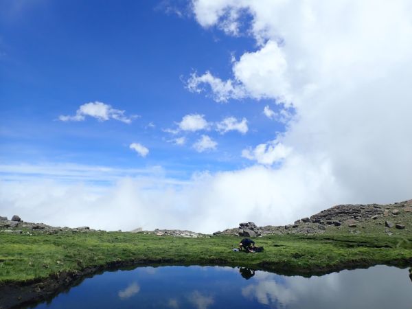 Man doing fieldwork next to a pristine lake, with a bright green grass bank against a vibrant sky.