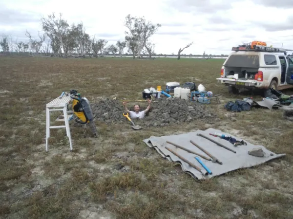 Fieldwork site with core samplers, equipment and vehicle. One man standing in a trench with hands in the air.