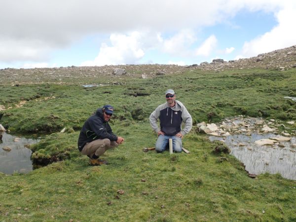 Two people working at a standpipe on a grassy spot next to a creek.