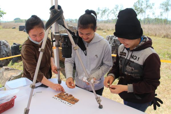 Three Laos University students standing by a table and a tripod cataloguing ceramic shards