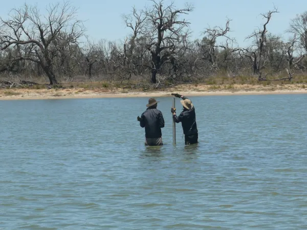 Two people stand in lack with core sampler. Trees without leaves are on the shore.