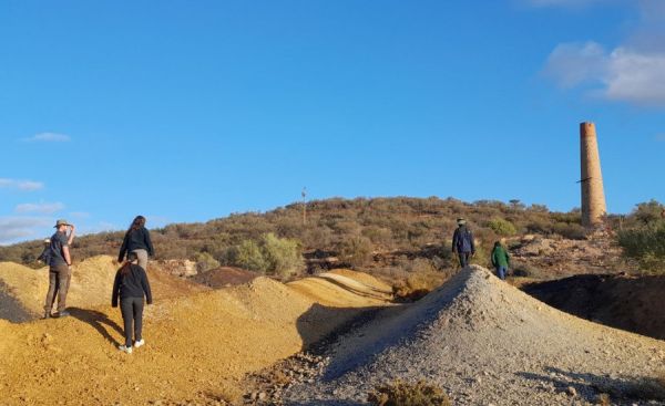 People walking across tailings headed up a scrubby hill toward a brick chimney.
