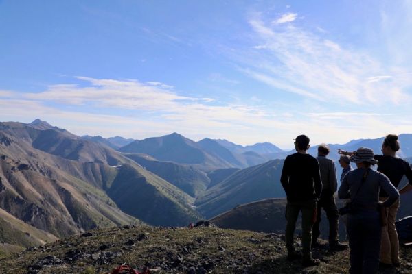 A rear view image of a group of researchers standing on a hill in Canada, looking out to a valley of green hills, with one researcher pointing to them.