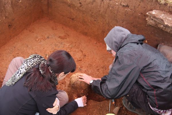 Two researchers, one with long brown hair and a spotted scarf, the other wearing a green raincoat, excavating a burial uni