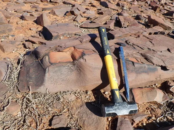 Ground covered in rust-coloured rocks with hammer and chisel leaning against a large rock.